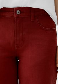 Tmavě červené džíny z denimu s texturovaným povrchem, obsahující přední kapsu a kovové zapínání na knoflík v pase.