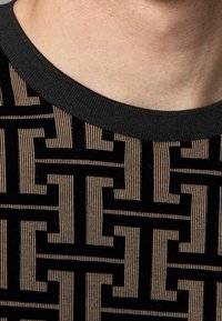 Suéter negro de manga corta con un patrón geométrico texturizado en tonos beige y crema, y un cuello redondo de canalé negro.
