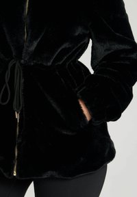 Giacca nera in pelliccia sintetica con vita regolabile tramite cordoncino, chiusura a zip e tasche laterali; presenta una texture morbida e eleganti dettagli in hardware dorato.