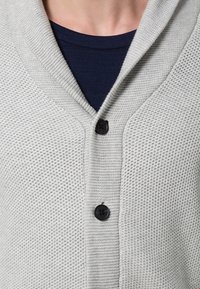 Světlounce šedý pletený cardigan s výstřihem do V, zdobený texturovaným vzorem a černými knoflíky. Nošený přes tmavě modrou košili.