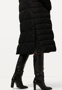 Abrigo acolchado negro con un patrón de rombos y botones laterales, combinado con botas de cuero negro brillante hasta la rodilla con tacones altos.