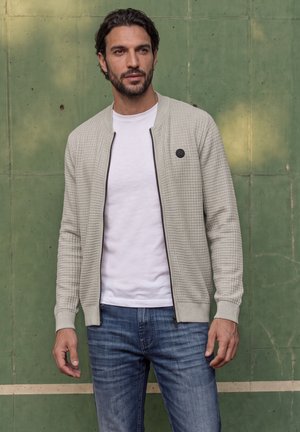 Un homme pose devant un mur vert, vêtu d’un cardigan gris clair zippé par-dessus une chemise blanche et d’un jean bleu.