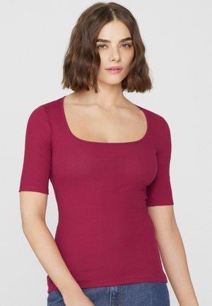 Vrouw met schouderlang golvend bruin haar die een aansluitend, geribd, kort mouw, vierkante hals bessenkleurig shirt en een blauwe jeans draagt.