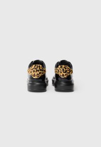Zwarte gepatenteerde leren sneakers met leeuwenprintaccenten op de hiel, gezien van achteren op een witte achtergrond.