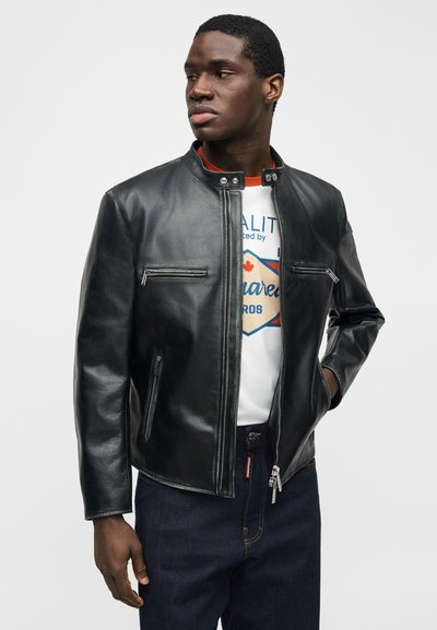 Νέος άνδρας που φοράει μαύρο δερμάτινο μπουφάν με φερμουάρ πάνω από λευκό μπλουζάκι με γραφικά και σκούρα μπλε τζιν, κοιτάζοντας προς τα αριστερά σε ένα απλό φόντο.