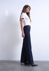 Νεαρή γυναίκα με μακριά μαλλιά, φορώντας λευκό μπλουζάκι, σκούρο τζιν με φαρδιά μπατζάκια, μαύρες ψηλοτάκουνες γόβες και ασημένια βραχιόλια, στέκεται πλάγια.