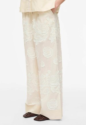 Kremfarvede bukser med vide ben og stor hvid blomsterbroderi, båret med brune vævede sko med spids tå og en matchende cremefarvet top.