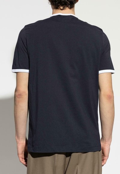 Hombre vistiendo una camiseta negra de manga corta con ribete blanco en las mangas y el cuello, combinada con pantalones marrones, visto desde atrás.