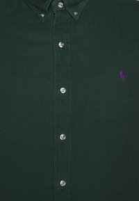 Tmavě zelená kordurová košile s límečkem na knoflíky, stříbrnými knoflíky a malým fialovým logem na hrudi. Texturovaná tkanina a přiléhavý střih.