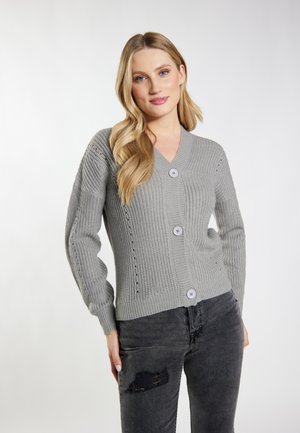 Femme aux longs cheveux blonds portant un cardigan en maille gris boutonné et un jean noir déchiré sur un fond clair uni.