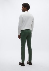 Λευκό πουκάμισο με μακριά μανίκια και στενά πράσινα παντελόνια. Τα παντελόνια διαθέτουν δύο πίσω τσέπες με διακριτικό εξάρτημα. Το μοντέλο φοράει σκούρα παπούτσια.