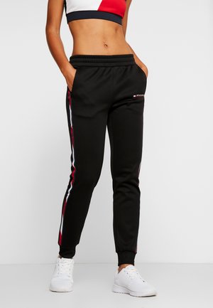 Frau trägt schwarze Jogginghosen mit roten und weißen Seitenstreifen, weiße Sneakers und ein rotes, weißes und schwarzes Crop-Top, steht mit den Händen in den Taschen.