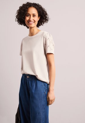 Femme souriante aux cheveux courts et bouclés portant un haut crème avec des manches courtes à œillets et un pantalon large en denim bleu, debout devant un fond clair.