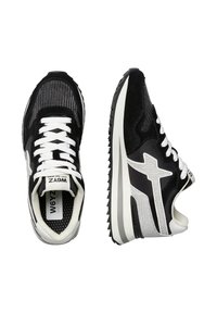 Sneakers bianco e nero con tomaia in rete e suede, caratterizzati da un pannello laterale con motivo, lacci bianchi spessi e una suola leggermente texturizzata.