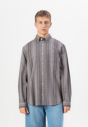 Young man wearing a long-sleeve buttoned plaid shirt and light blue jeans, standing against a plain white background.