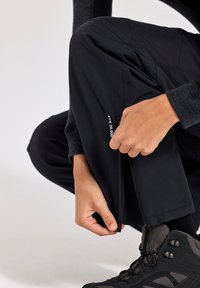 Schwarze Outdoor-Hosen mit Reißverschluss am Knöchel, aus weichem Material und einem wasserdichten Etikett. Getragen mit strukturierten schwarzen Wanderschuhen.