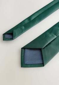 Sangles en tissu vert avec une surface texturée, présentant un intérieur à pois bleus. Les extrémités sont de forme carrée et soigneusement finies.