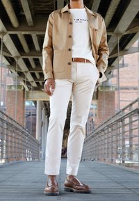 Americana beige con botones sobre una camiseta blanca, pantalones crema claros y mocasines marrones. Fondo industrial con suelo de madera y barandillas.
