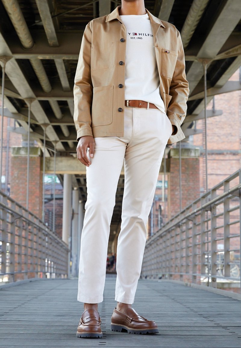 Americana beige con botones sobre una camiseta blanca, pantalones crema claros y mocasines marrones. Fondo industrial con suelo de madera y barandillas.