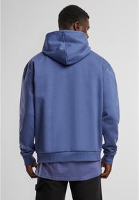 Felpa con cappuccio blu realizzata in tessuto morbido, con una vestibilità comoda, polsini e orlo a costine, e una tasca a marsupio. Indossata sopra una maglietta viola.