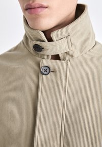 Beige kordfløyelsjakke med høyt krage, med svarte knapper og en ribbestruktur. Nærbilde viser detaljene på kragen og plasseringen av knappene.