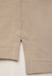 Beige textiel met structuur, met een shirtzoom en een zijsplit; stiksels zijn zichtbaar en voegen structuur toe aan het ontwerp.