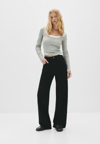 Femme blonde portant un crop top gris ajusté à manches longues, un pantalon noir large taille haute et des chaussures foncées, debout avec une main dans la poche.