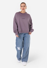 Felpa viola con colletto a giro e polsini e orlo a costine, abbinata a jeans azzurri a vita alta con dettagli strappati sulle ginocchia. Sneakers bianche.