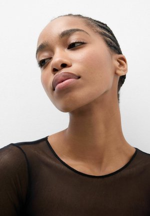 Joven con trenzas y una blusa negra transparente, mirando ligeramente hacia un lado sobre un fondo blanco liso.
