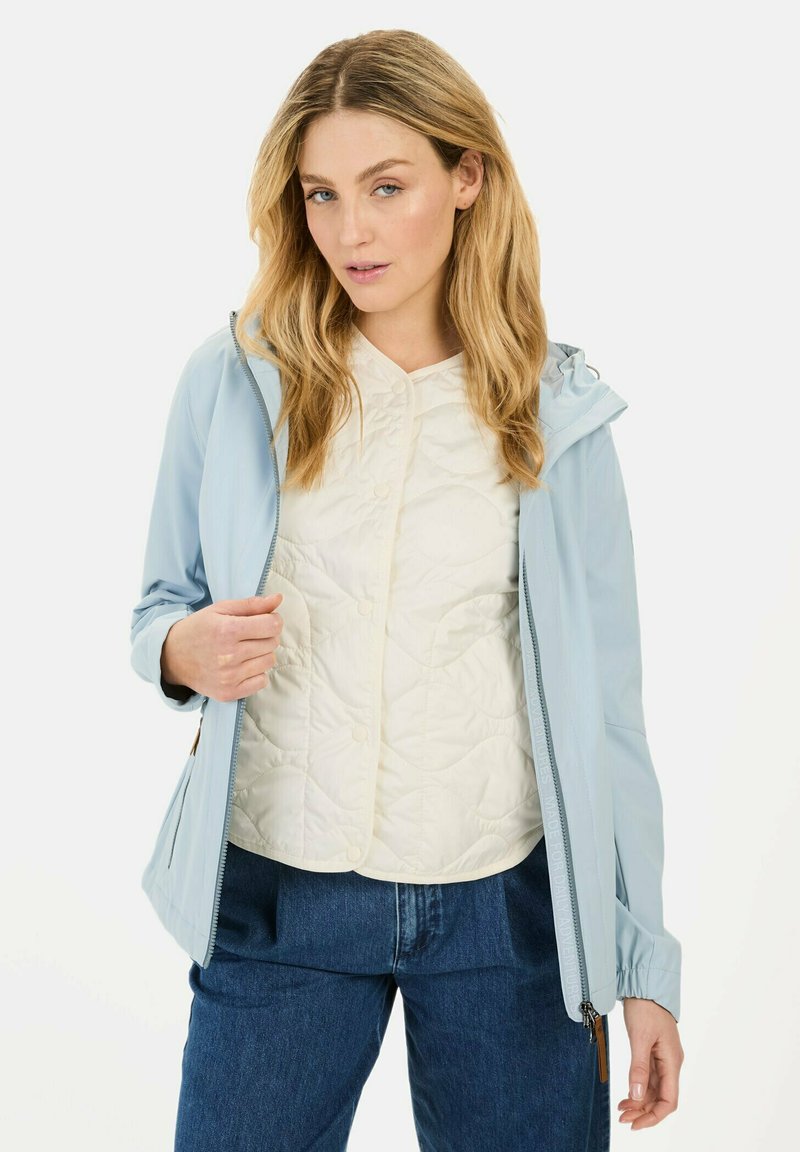 Lysblå jakke med hætte, lavet af glat materiale. Båret over en hvid quiltet bluse med rund halsudskæring og knapper. Mørke denim bukser.