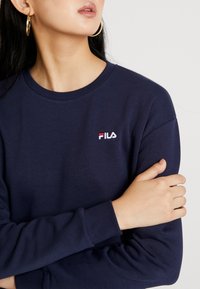 Tmavě modrá mikina s dlouhým rukávem a kulatým výstřihem, na hrudi vyšitý bílý a červený nápis FILA. Měkká textura.