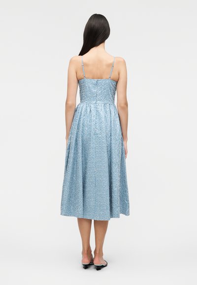 Γυναίκα με μακριά σκουρόχρωμα μαλλιά, που φοράει μπλε υφασμάτινο μίντι φόρεμα χωρίς μανίκια με λεπτές τιράντες, στέκεται με την πλάτη σε απλό φόντο.