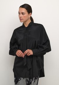 Juoda satino blusa su aukštu apykakle ir kaklo juostele. Ilgos rankovės, susirinkusios ties riešais. Laisvas kirpimas su šiek tiek išlenktu apvadu.