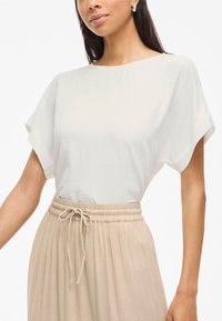 Blusa bianca a maniche corte con una texture liscia, scollatura rotonda e vestibilità ampia. Abbinata a una gonna beige a vita alta con coulisse.