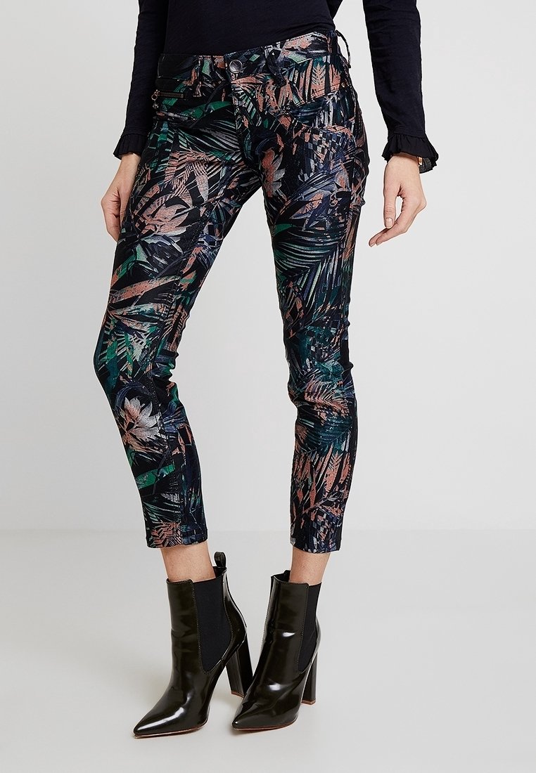 Femme portant des bottines noires et un pantalon ajusté avec un motif coloré de feuilles tropicales, debout devant un fond uni.