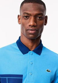 Turkos polo skjorta med marinblå krage, med ett grafiskt mönster och Lacoste-logotyp som accent. Mjuk textur och knäppdetaljer synliga.
