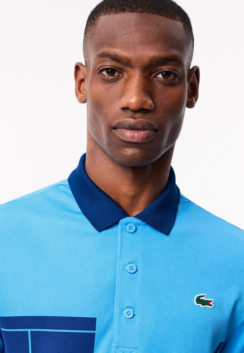 Polo turquoise avec un col marine, présentant un motif graphique et un accent du logo Lacoste. Texture douce et détails de fermeture à boutons visibles.