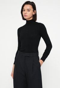 Čierny top s dlhým rukávom a rolákom, priliehavého strihu, vyrobený z hladkej látky, v kombinácii s tmavými nohavicami s jemným pásikovým vzorom, ruky vreckách.