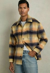 Chaqueta de camisa a cuadros en tonos amarillo, azul y negro. Cuenta con dos bolsillos frontales, cierre de botones y un acabado de tela texturizada.