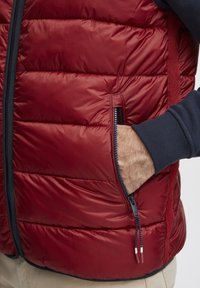 Veste doudoune rouge avec un quilting horizontal, dotée d'une poche zippée, d'un parement bleu marine et de poignets élastiques. Texture lisse avec une finition brillante.