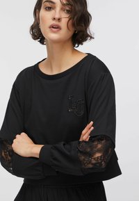 Sweat-shirt noir à manches longues avec détails en dentelle, encolure ronde et logo brodé. Matière douce avec coupe décontractée.