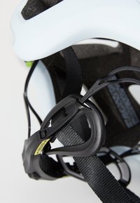 Gros plan sur un casque bleu clair avec des sangles noires ajustables et un clip de réglage vert, mettant en valeur son design intérieur et son système de fixation.