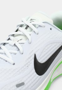 Αθλητικό παπούτσι ανοιχτού γκρι με μαύρο λογότυπο Nike, υφή πάνω επιφάνειας, λευκά κορδόνια και πράσινες λεπτομέρειες. Διαθέτει μαλακή σόλα για άνεση.