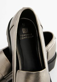 Mocasines plateados metálicos con un acabado texturizado, interior negro y logo de la marca elevado. Punta redondeada y suela baja y resistente de color negro.