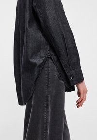 Μαύρο τζιν πουκάμισο με μακριά μανίκια, πλάγιες σχισμές και κουμπιά στους καρπούς, σε συνδυασμό με ταιριαστά σκούρα τζιν παντελόνια, παρουσιάζοντας μια συνεκτική εμφάνιση.