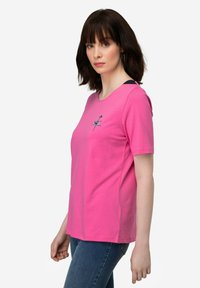 Roze katoenen T-shirt met korte mouwen, ronde hals en geborduurd ontwerp aan de voorkant. Gecombineerd met blauwe spijkerbroek.