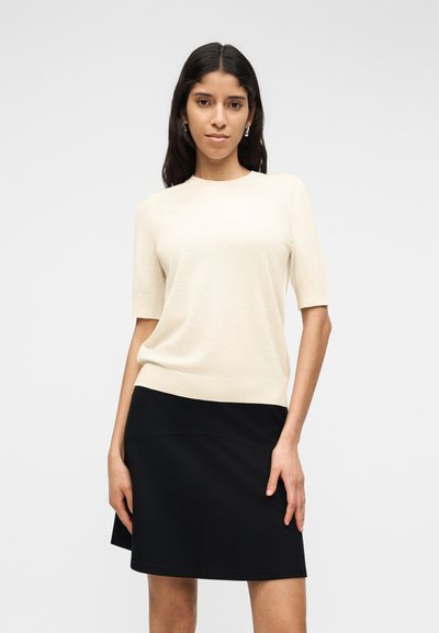 Femme aux longs cheveux bruns portant un haut crème à manches courtes et une jupe noire longueur genoux, debout devant un fond uni clair.