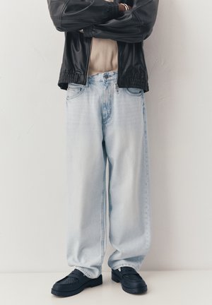 Person iført en sort læderjakke, beige skjorte, lyseblå løstsiddende jeans og sorte loafers, stående med krydsede arme mod en hvid væg.