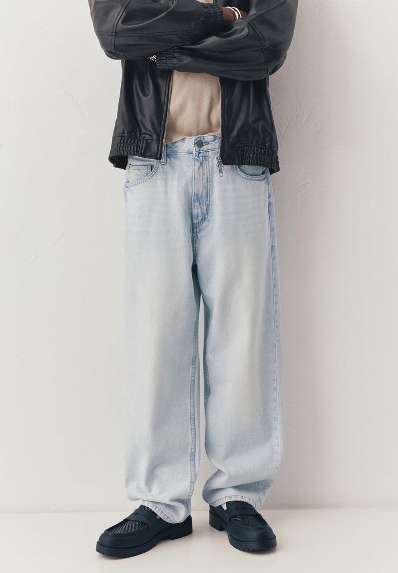 Personne portant une veste en cuir noire, une chemise beige, un jean bleu clair ample et des mocassins noirs, debout les bras croisés contre un mur blanc.