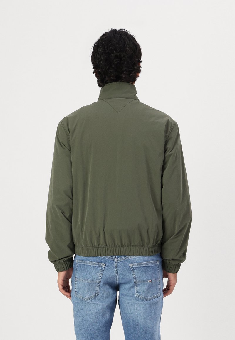 Veste coupe-vent vert olive avec un col haut, des poignets élastiques et une taille élastique. Texture lisse et design minimaliste.